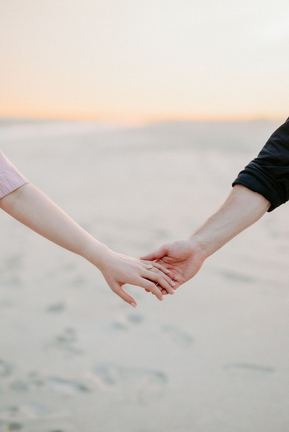 Couple holding hands on a beach at sunset, woman wearing an engagement ring, romantic outdoor moment.