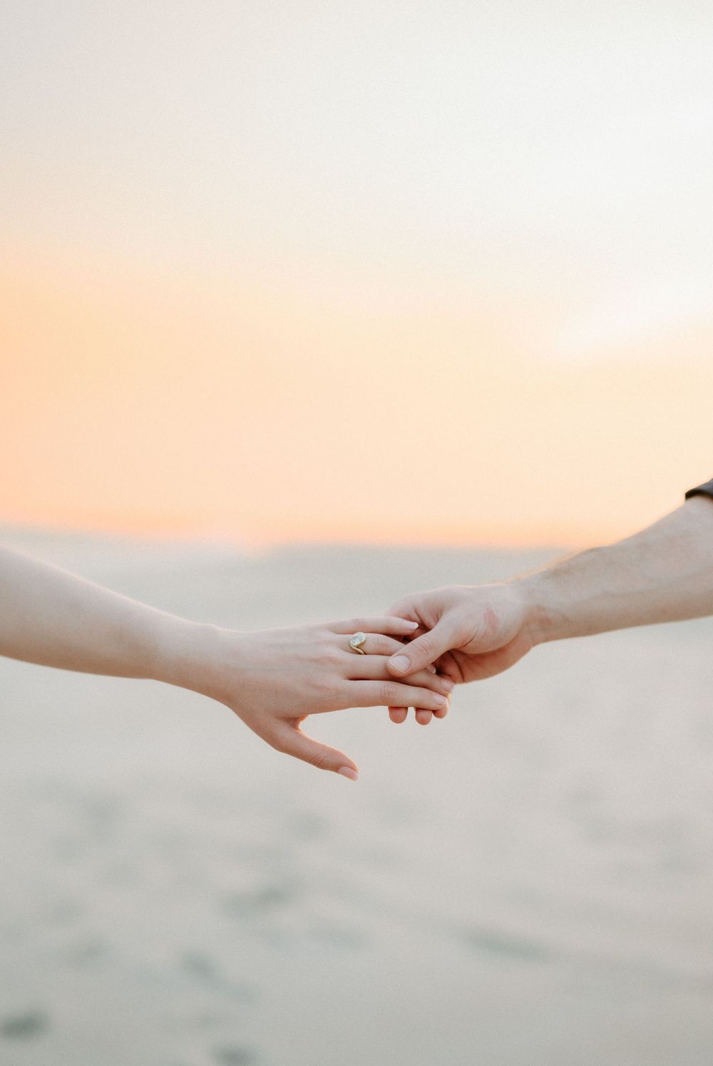 Two hands gently touching at sunset on a beach, with one hand wearing a gold engagement ring.