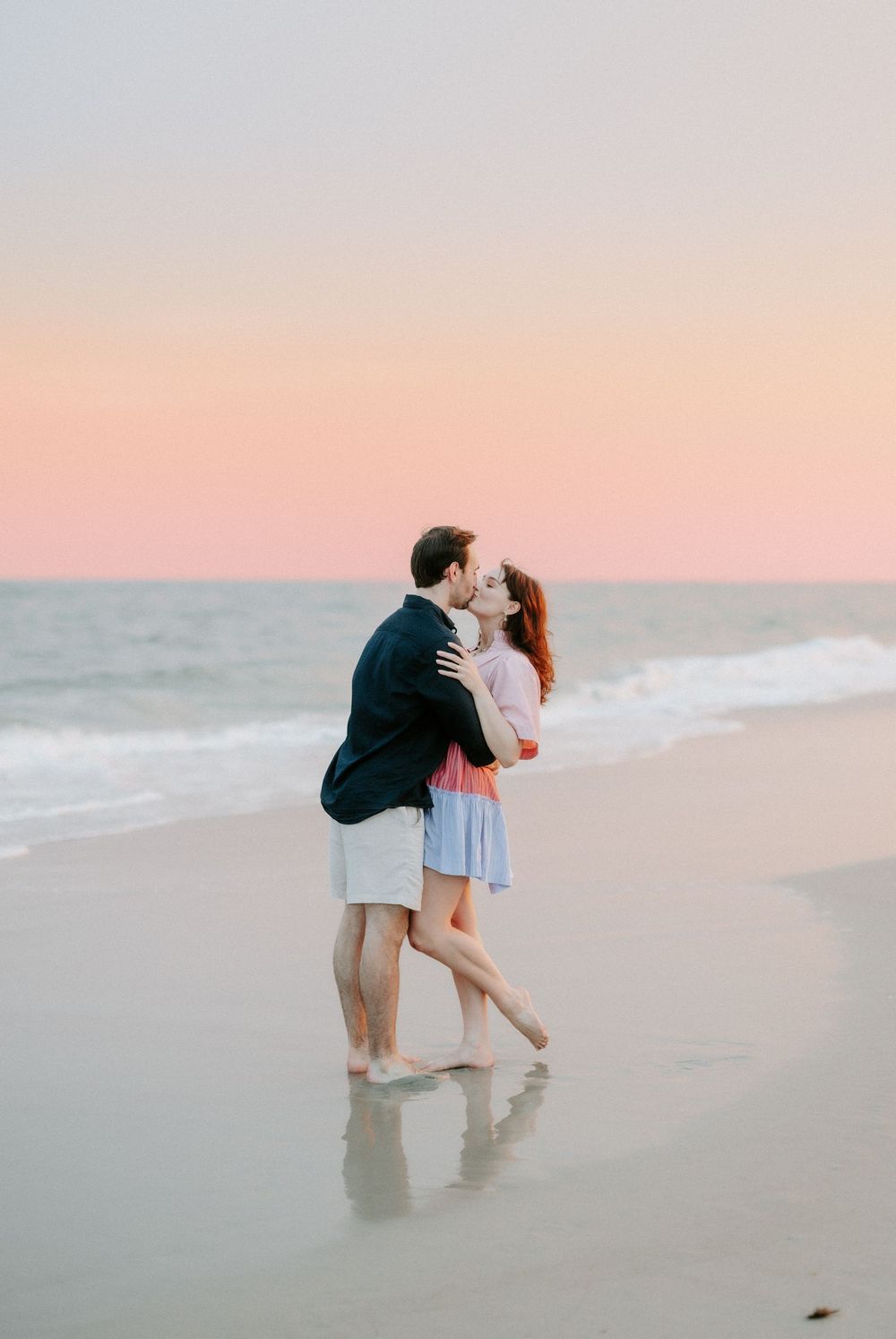 Couple sharing a romantic kiss on a sandy beach at sunset with soft pink sky and ocean waves.