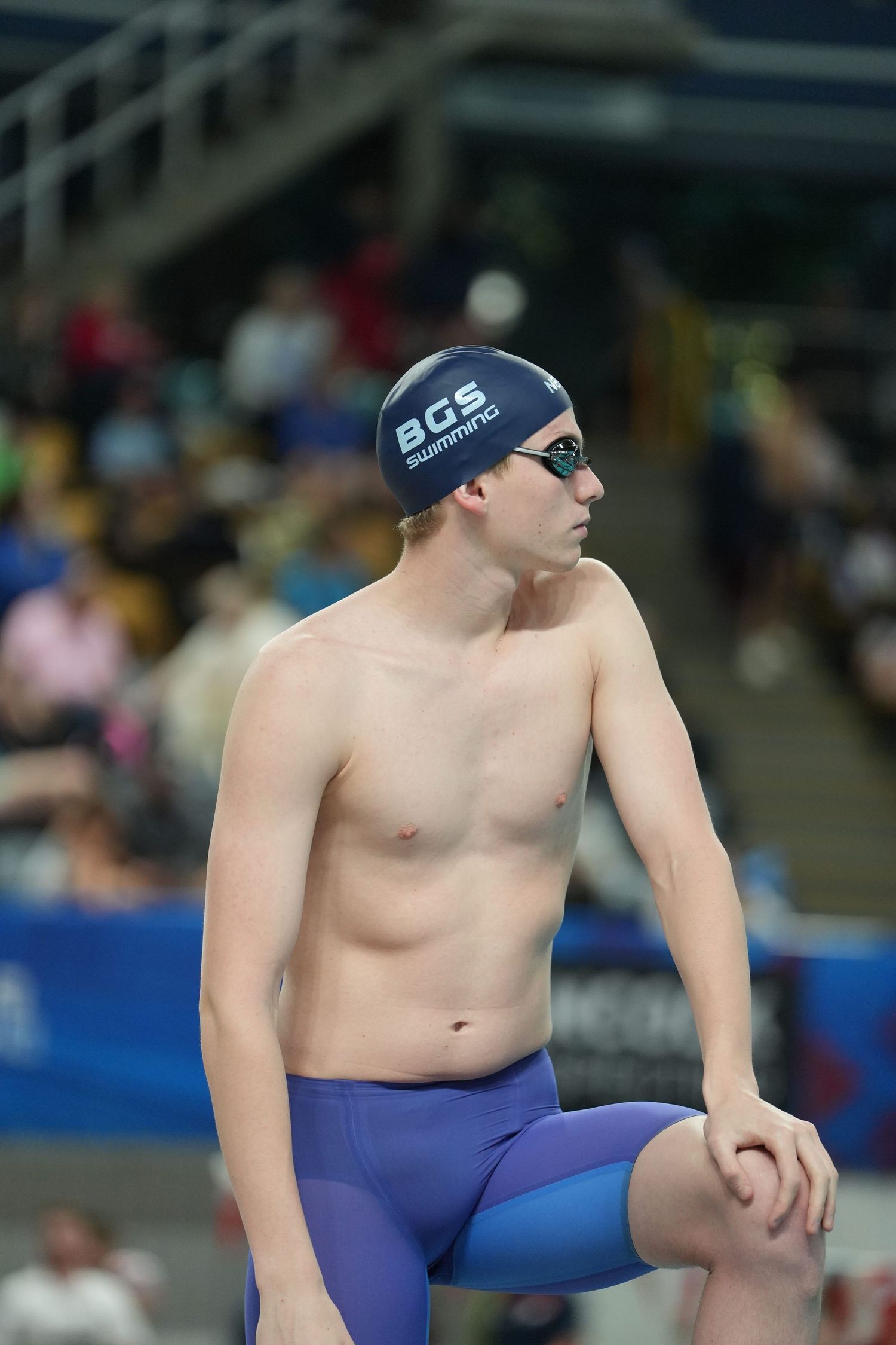 Young male swimmer in BGS Swimming cap and goggles stands poolside in competitive swimwear at an indoor aquatic event.