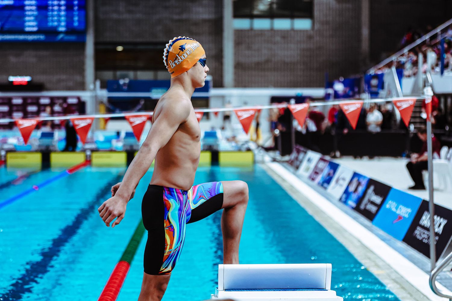 Young male swimmer in BGS Swimming cap and goggles stands poolside in competitive swimwear at an indoor aquatic event.