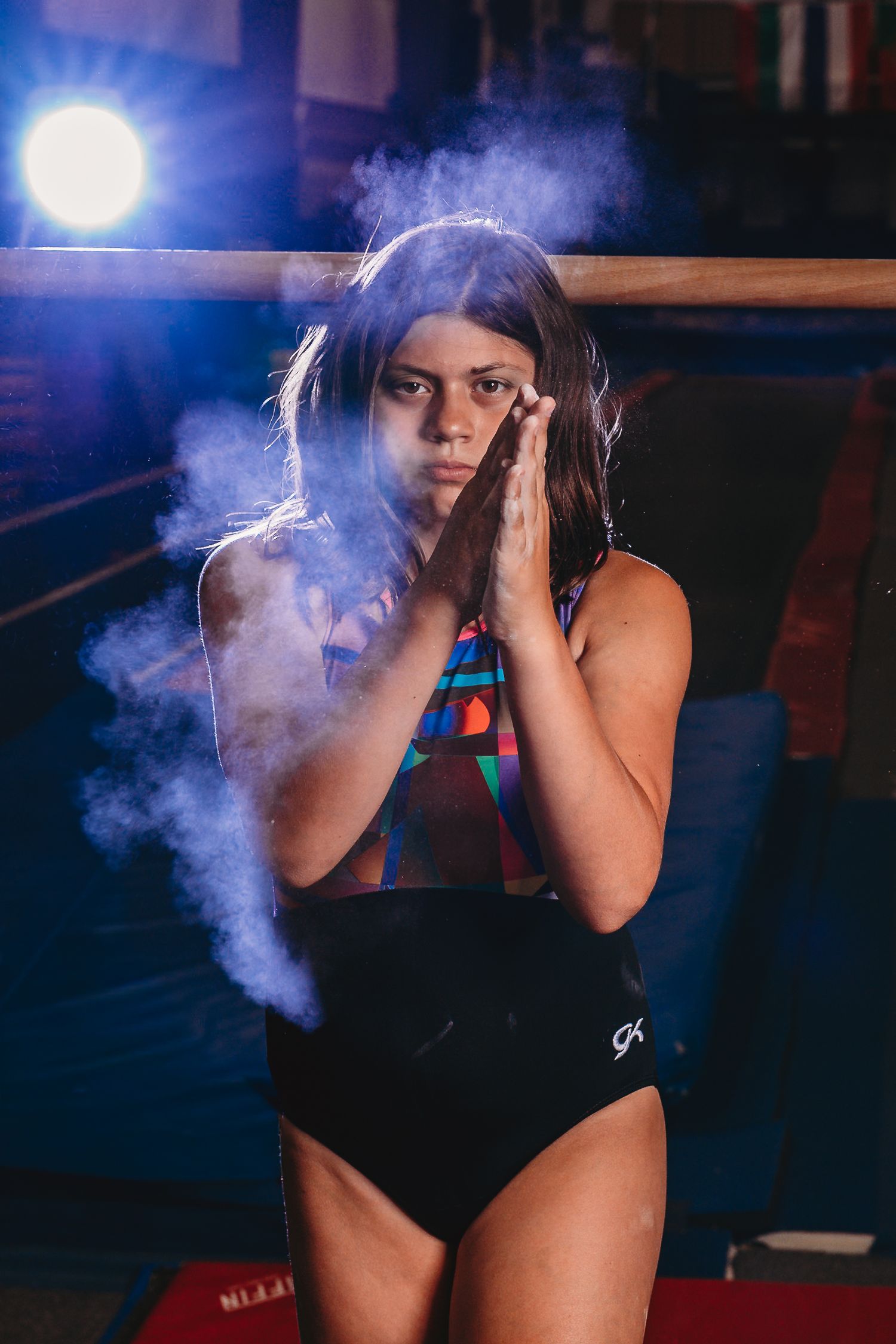 Female gymnast in GK leotard clapping chalk from hands in dramatic gym lighting.
