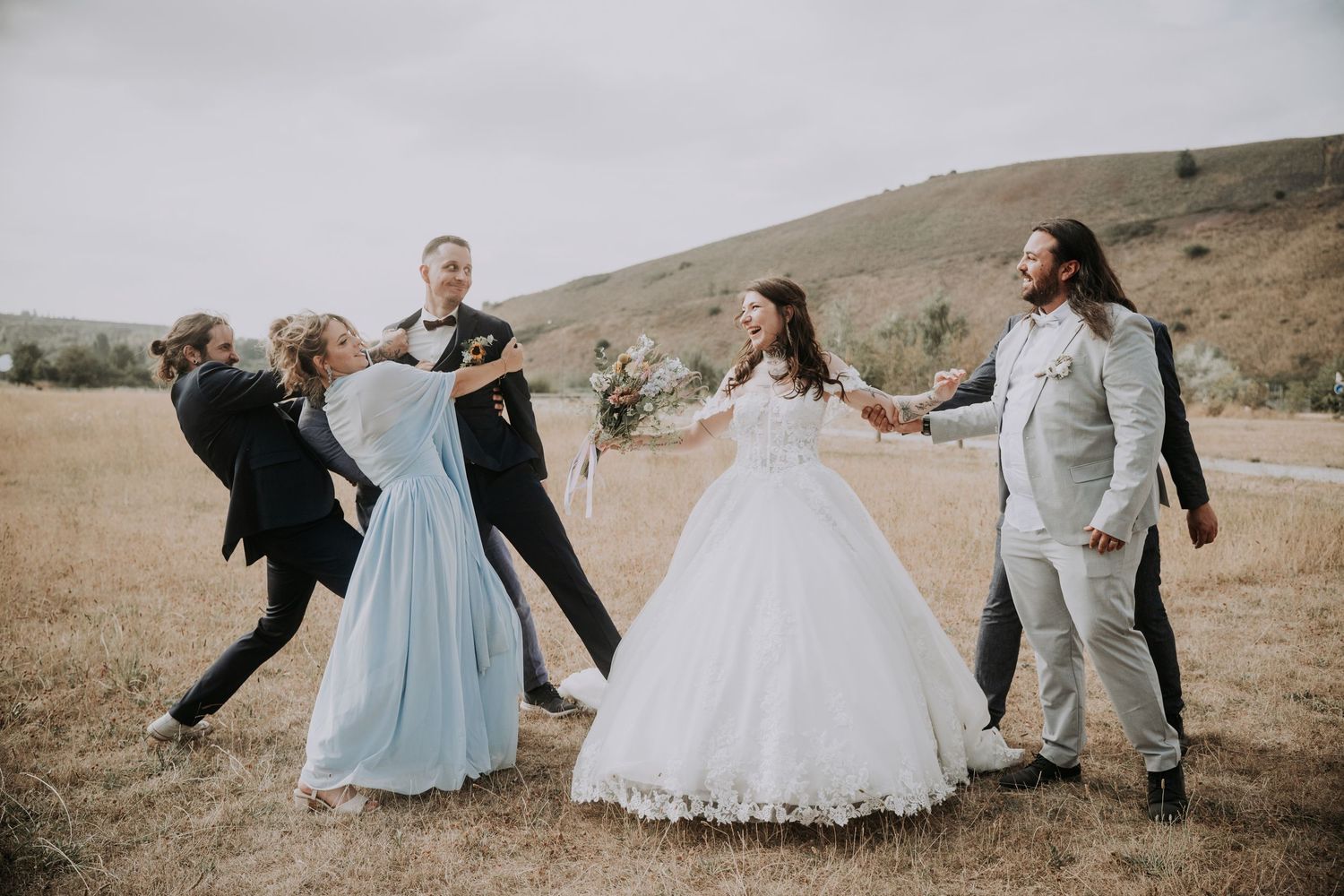 Wedding party celebrates outdoors in a field, bride in white gown holds hands with guests in a joyful circle dance.