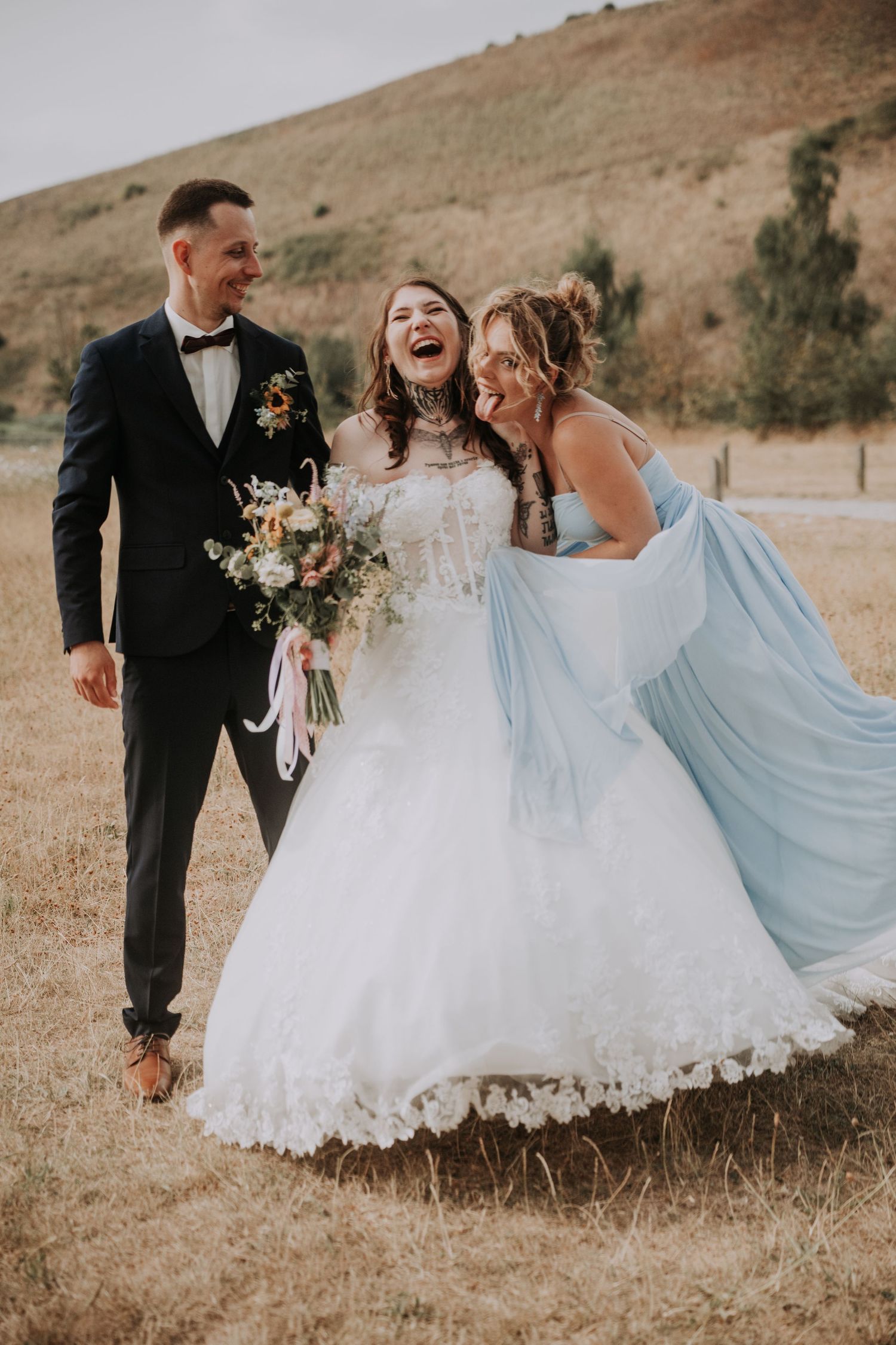 Bride in white gown laughing with groom in navy tuxedo and bridesmaid in light blue dress outdoors.