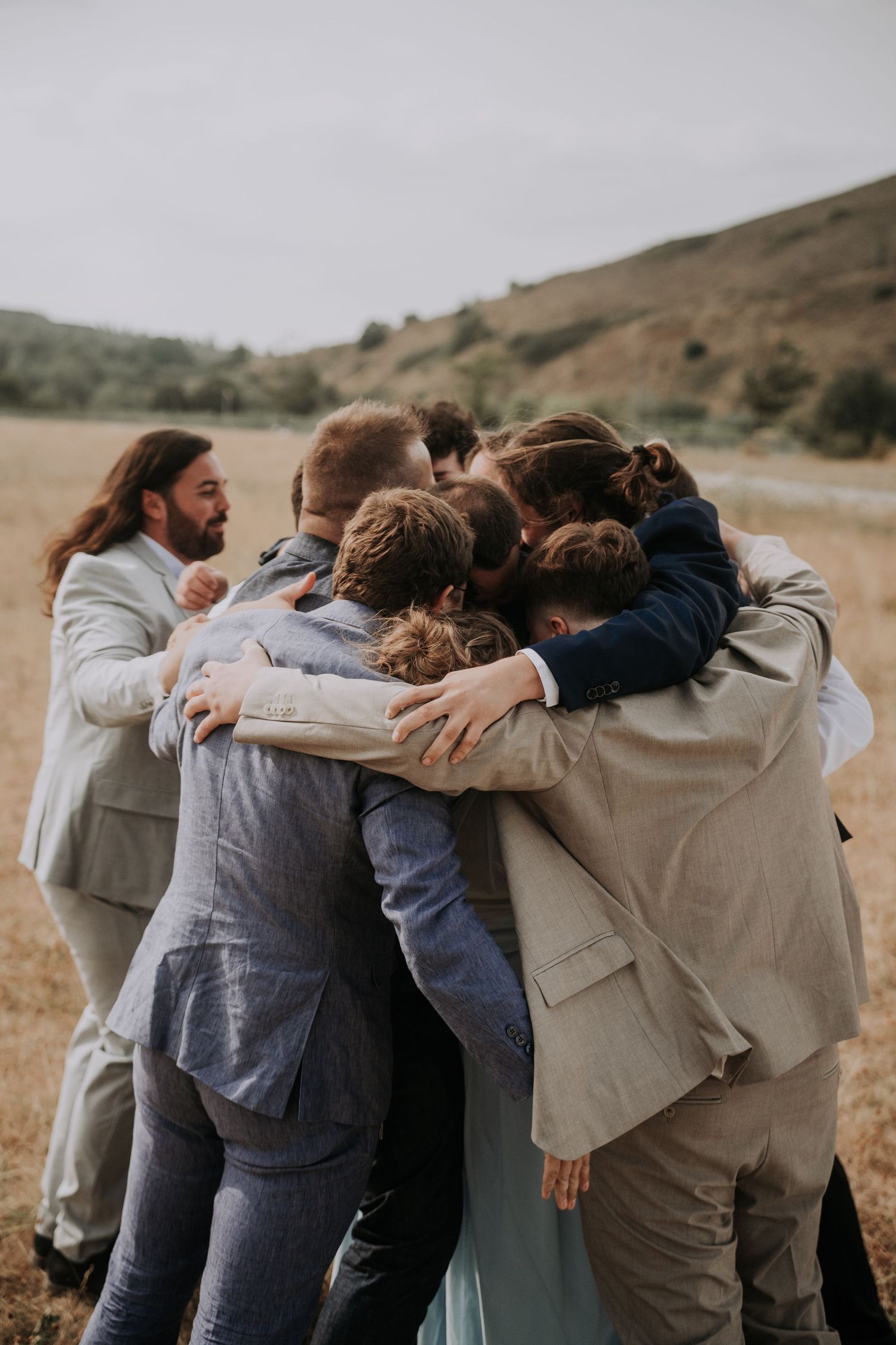 A group of wedding guests in formal attire share a warm group hug in an open field with rolling hills.