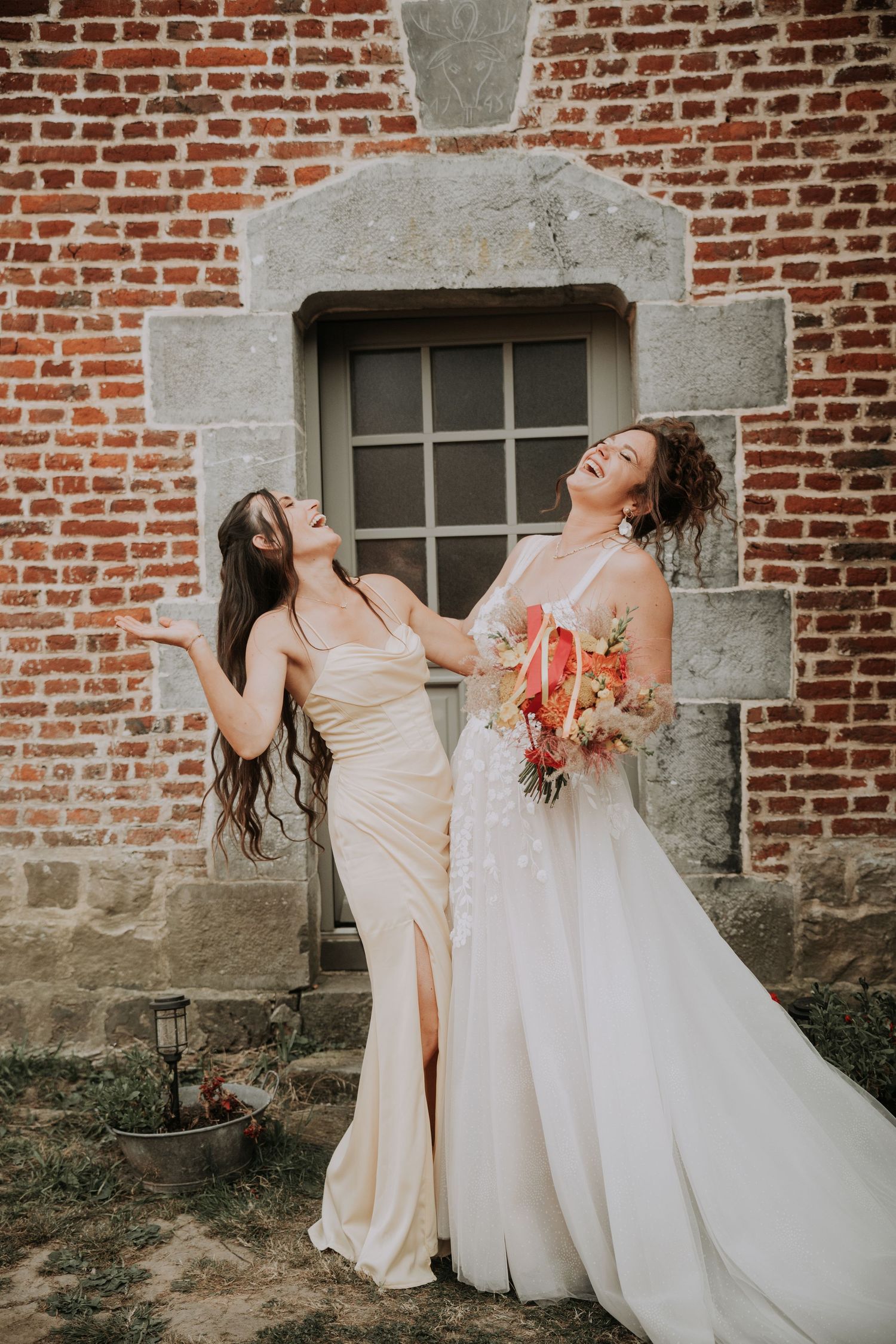 Two brides laughing and posing in elegant gowns, holding a colorful bouquet against a rustic brick wall.