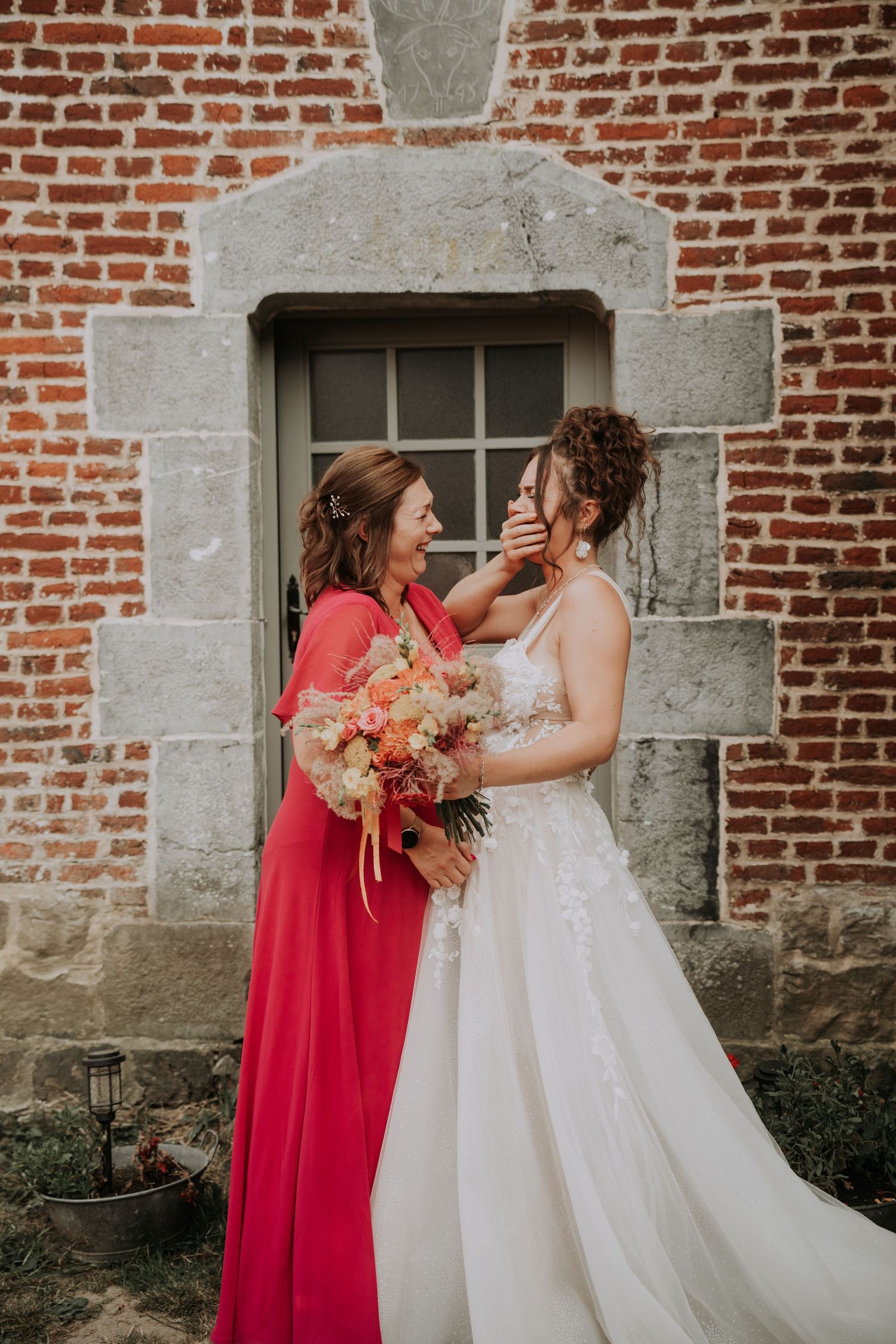Bride in white gown and woman in red dress share emotional moment outside rustic brick building with bouquet.