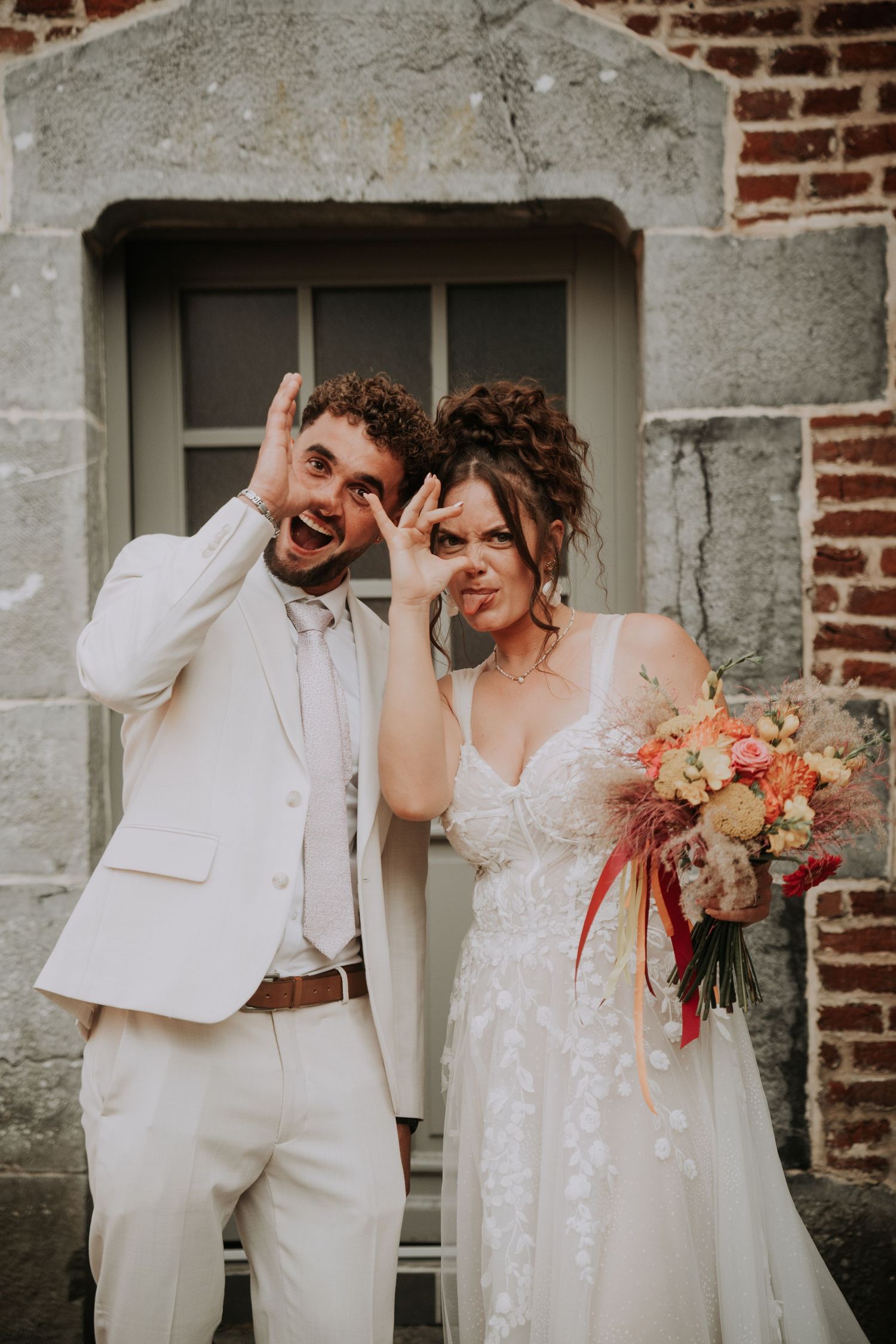 Playful bride and groom making fun gestures on their wedding day, bride holding colorful bouquet outside stone building.