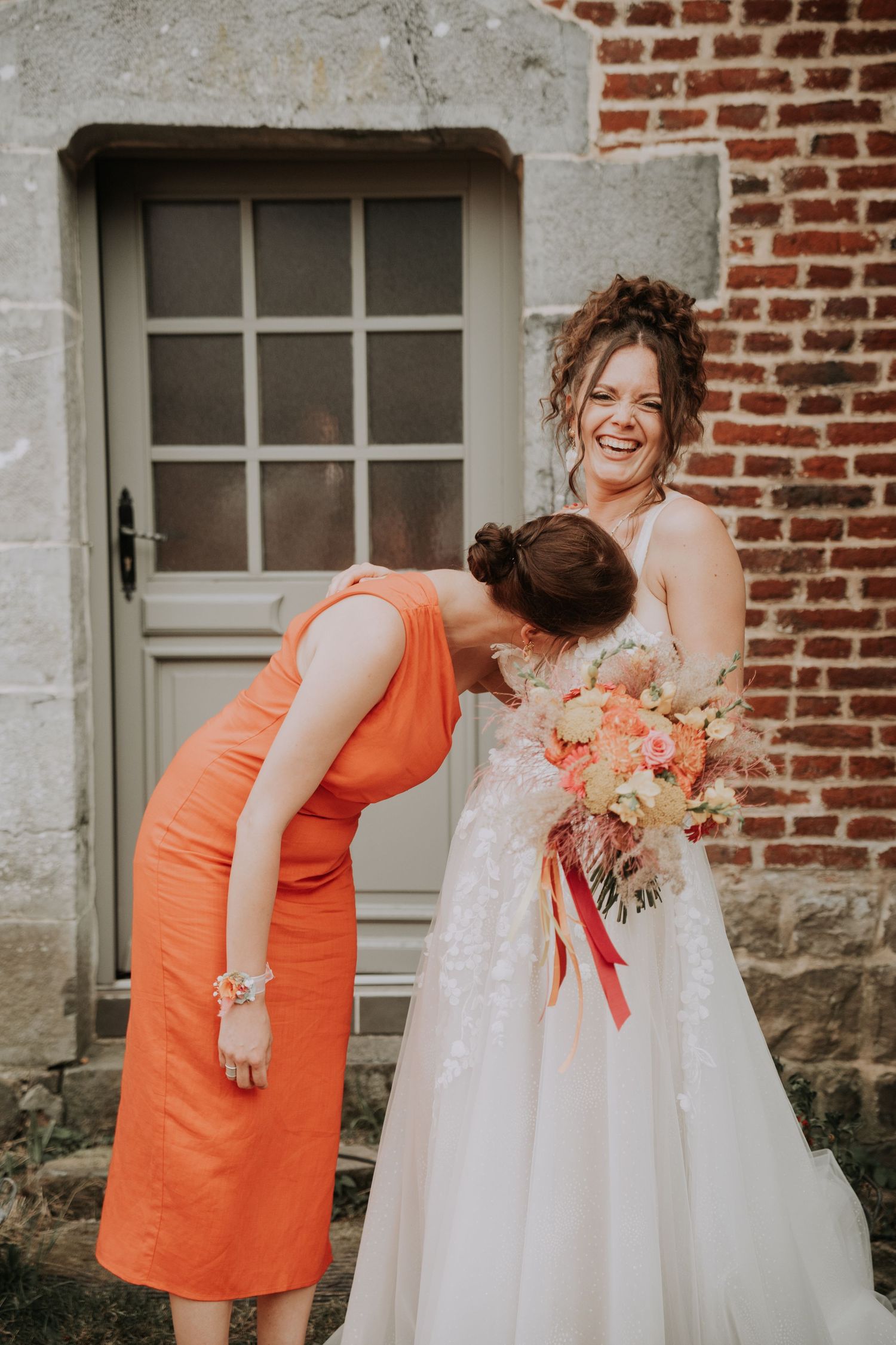 Bride in white gown laughs joyfully as bridesmaid in orange dress kisses her shoulder, holding colorful bouquet.