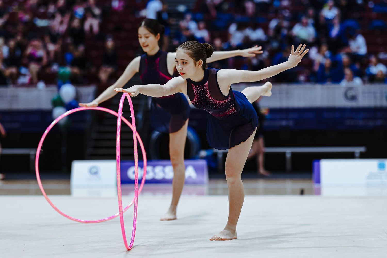 Two rhythmic gymnasts perform synchronized routine with pink hoops on competition floor in matching navy uniforms.