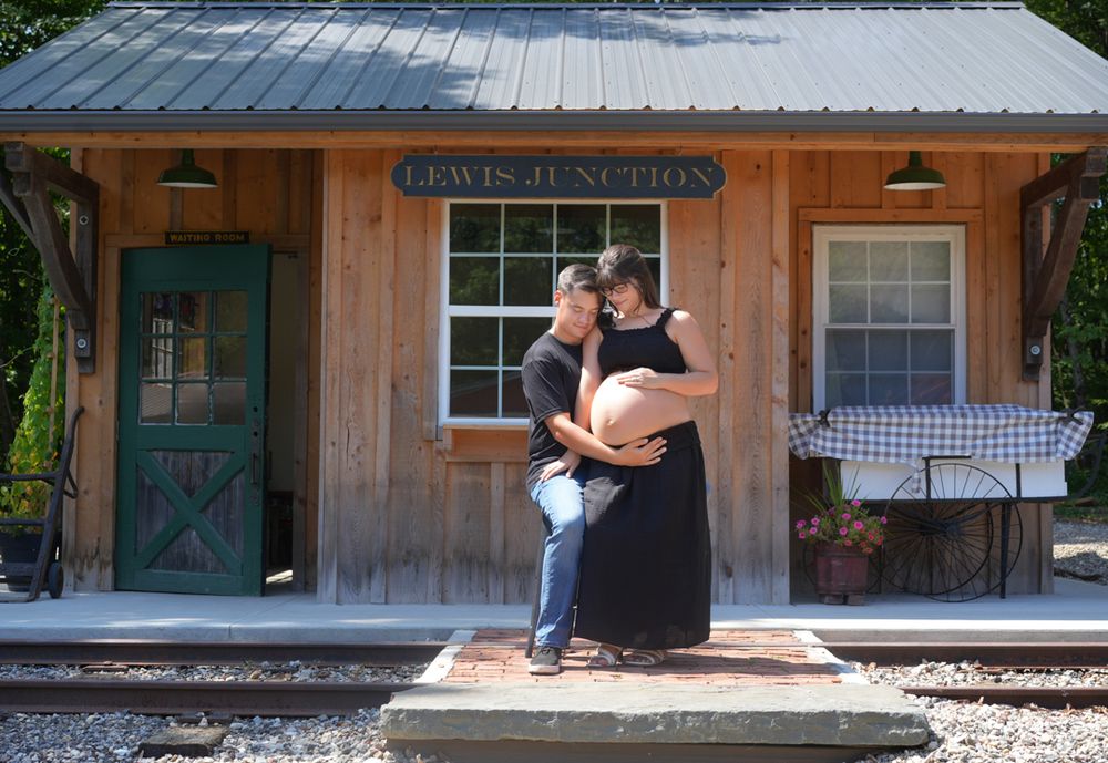 A couple embraces affectionately on a rustic wooden porch in front of a station building with a green door.
