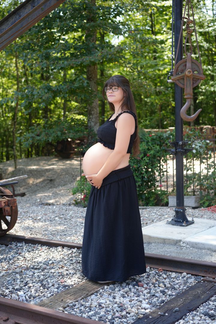 A pregnant woman in a black maxi dress stands on train tracks surrounded by stones and greenery in a peaceful outdoor setting.