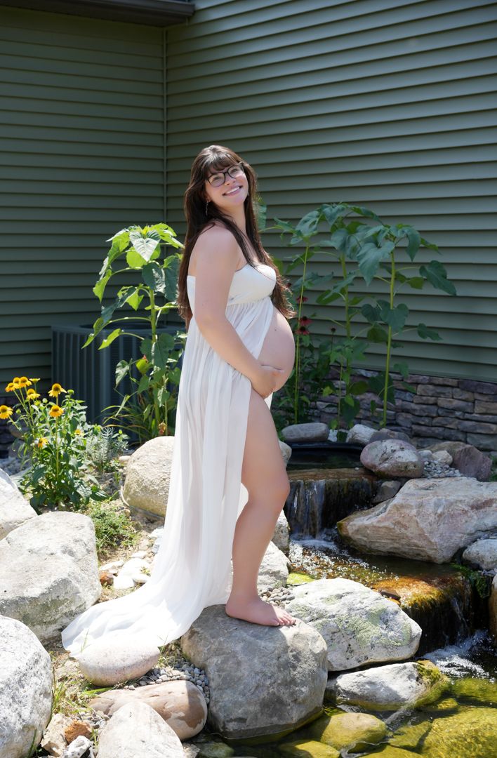 A pregnant woman in a flowing white dress poses barefoot on rocks near a garden water feature and green siding.