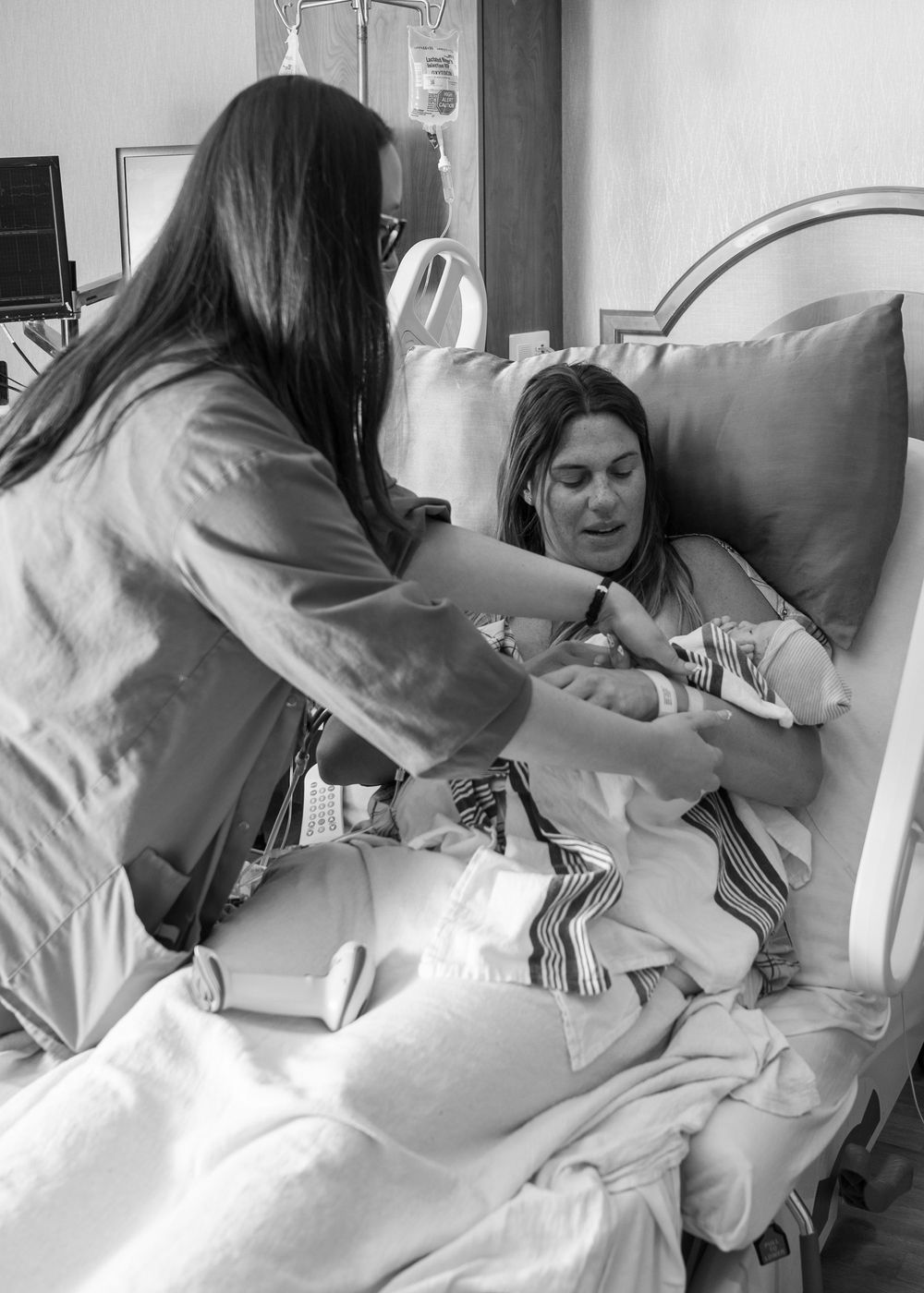 Black and white photo of a new mother in hospital bed holding her newborn baby with a nurse assisting.