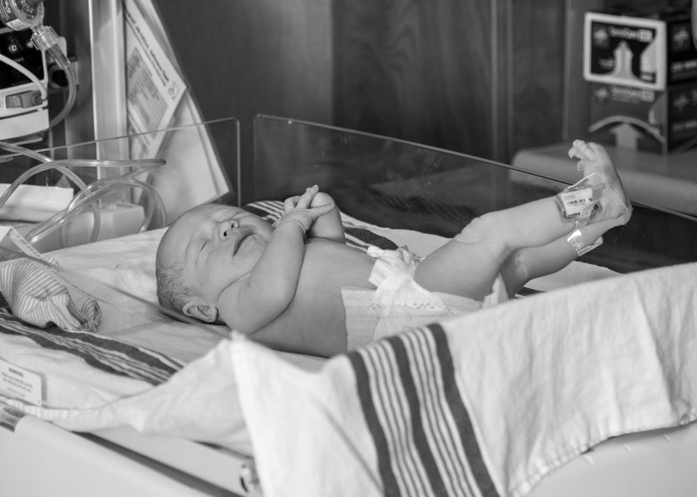 Black and white photo of a newborn baby lying in a hospital bassinet with medical monitoring equipment nearby.