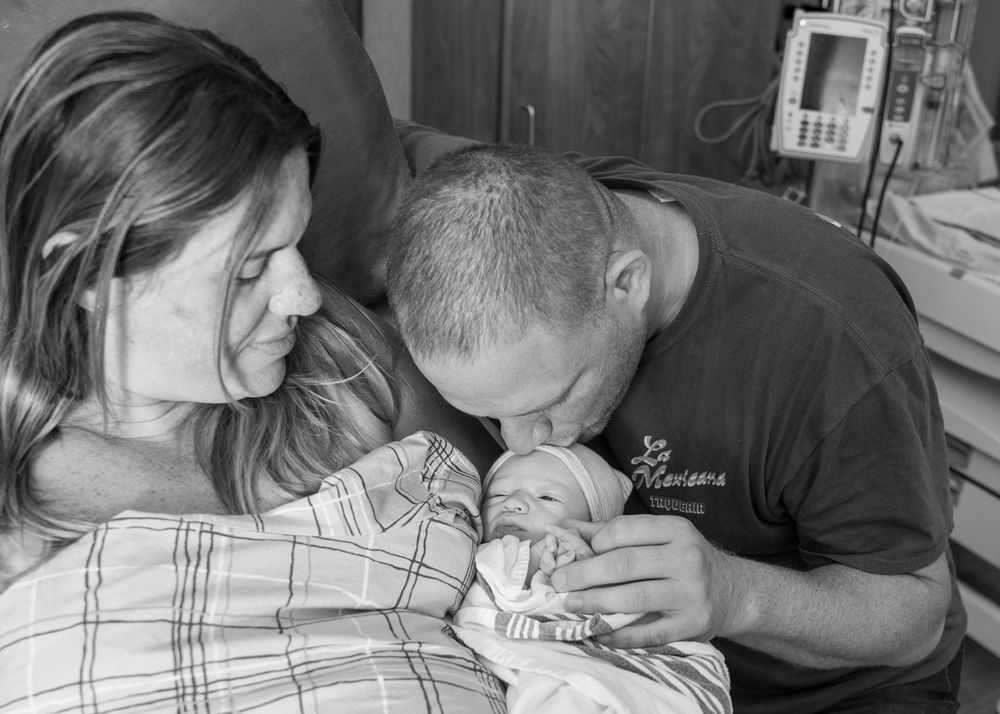 Black and white photo of new parents lovingly gazing at their newborn baby in a hospital room.
