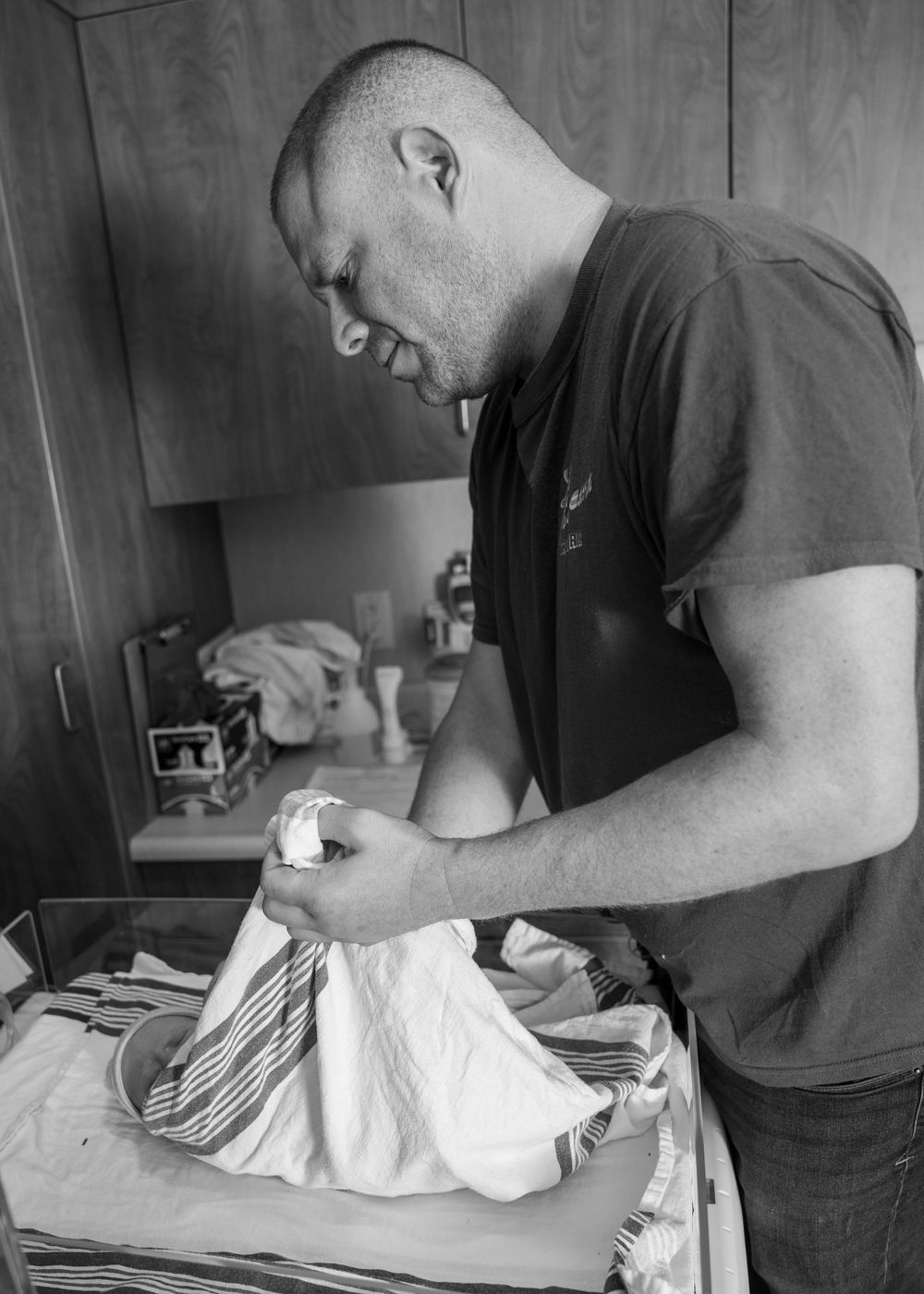 Black and white photo of a man folding laundry on a bed near a wooden wardrobe.