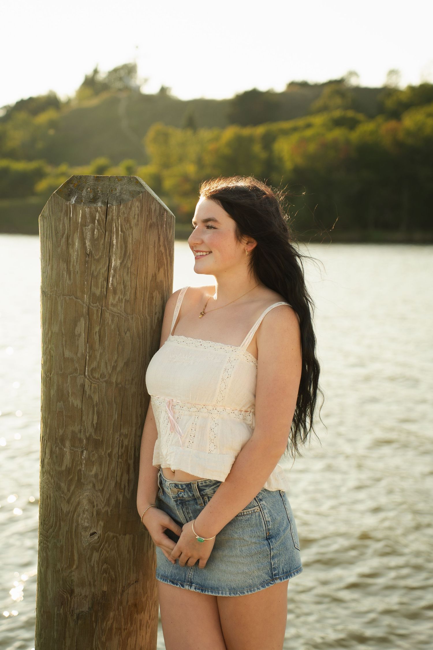 Young woman in white lace crop top and denim skirt smiling by a wooden dock post with sunlit water behind her.