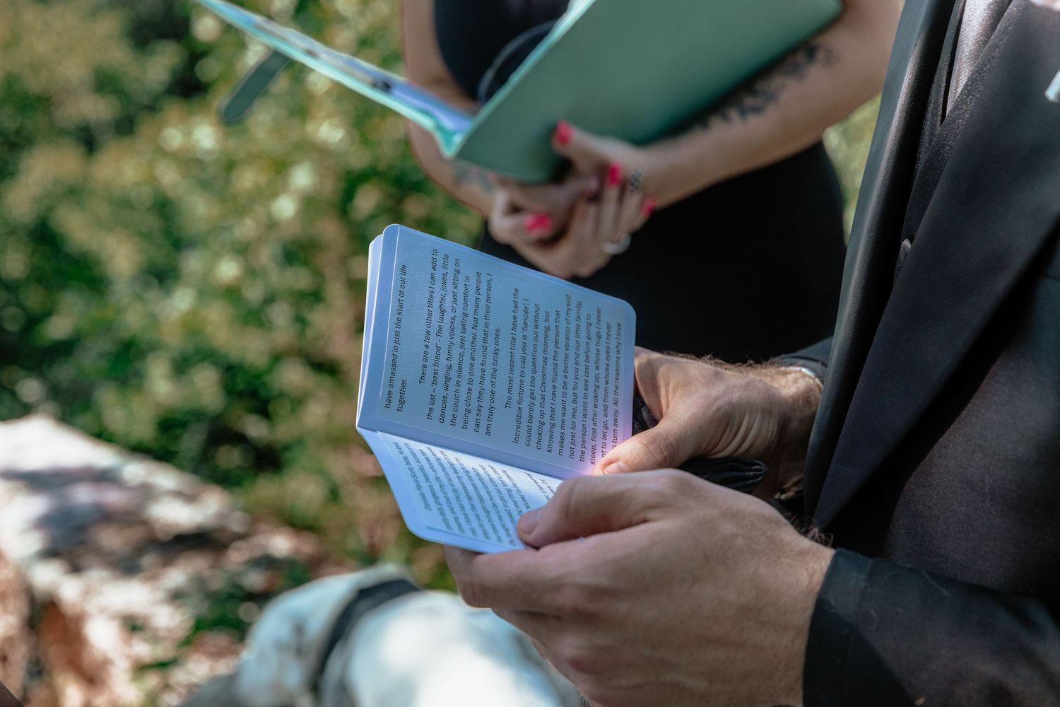 Two people holding small books outdoors, one with red nails and a tattoo, reading during a ceremony.
