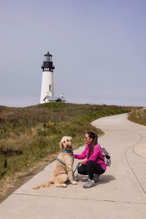 Woman in pink jacket crouching with golden doodle dog on path near white lighthouse on grassy hill.