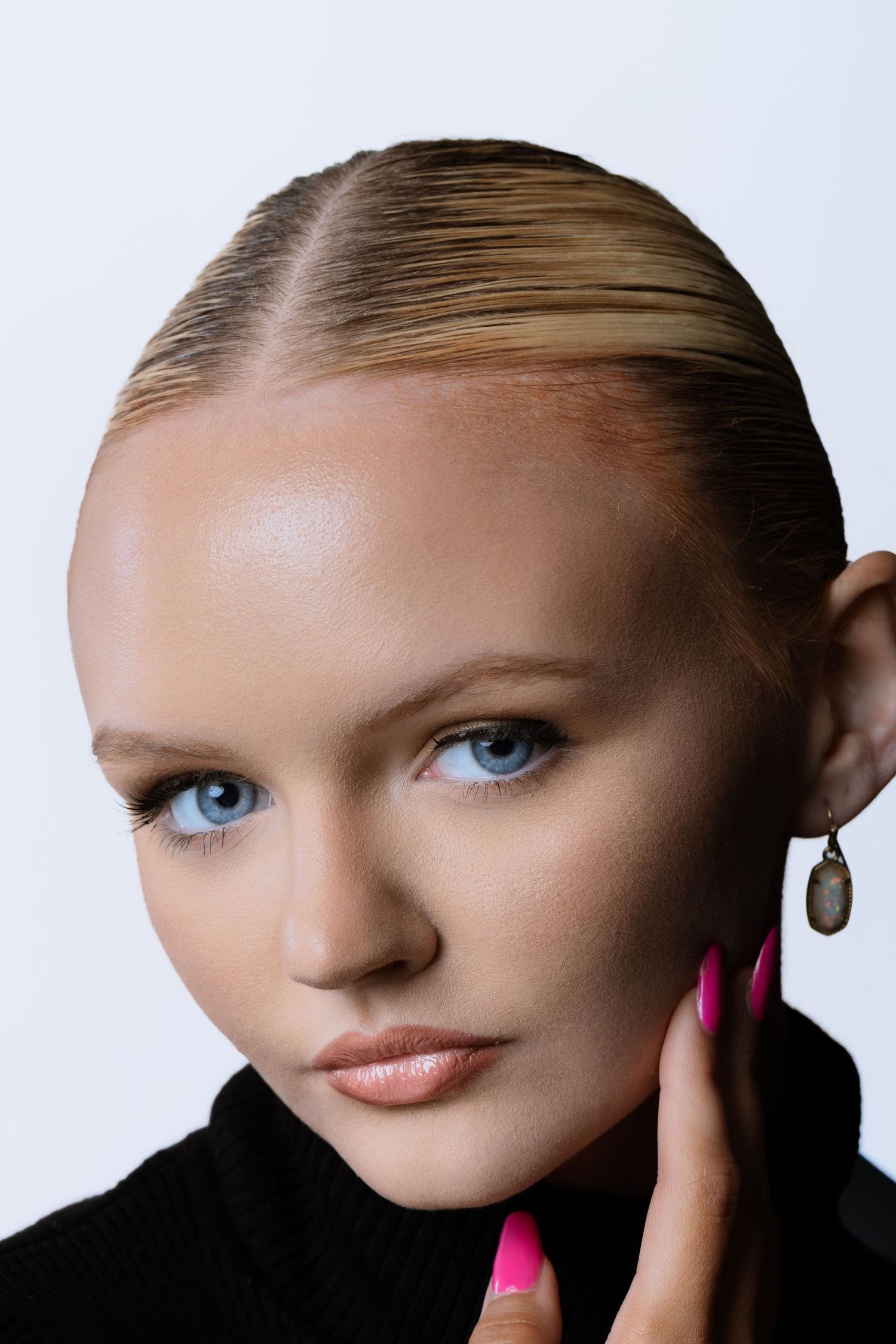 Close-up of a blonde model with blue eyes, slicked-back hair, drop earring, and bright pink nails against a white background.