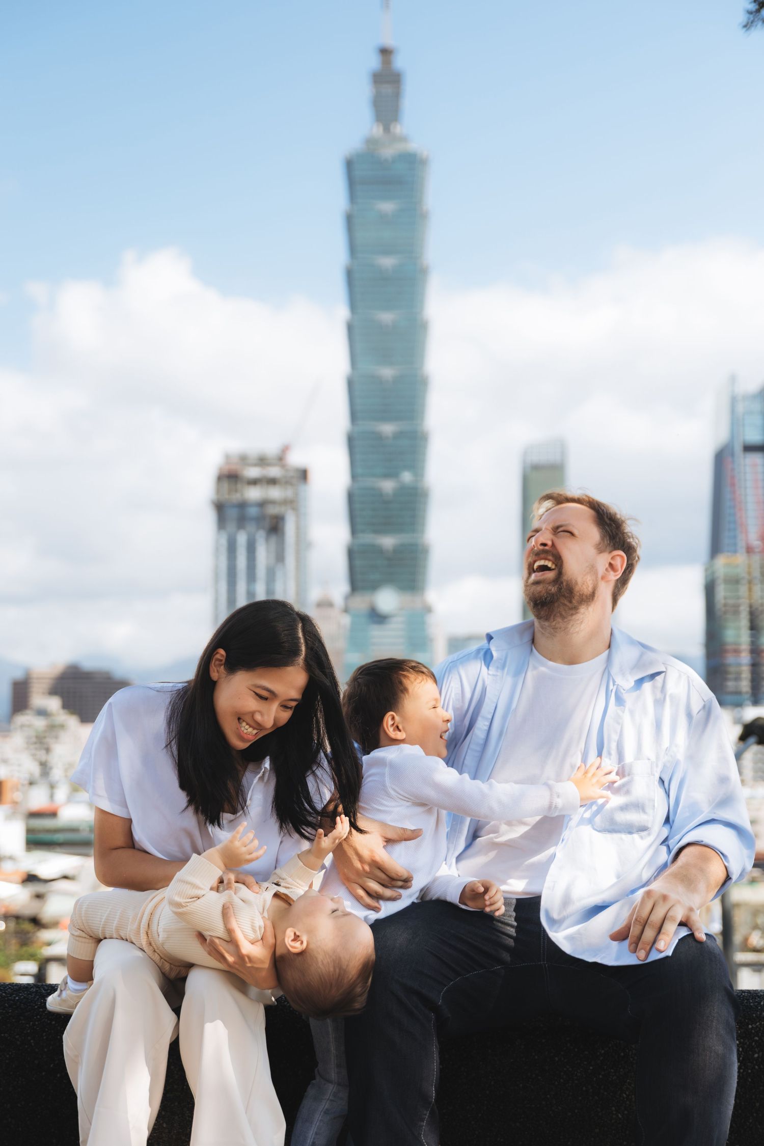 Happy mixed family with two young children posing outdoors with Taipei 101 skyscraper in the background.