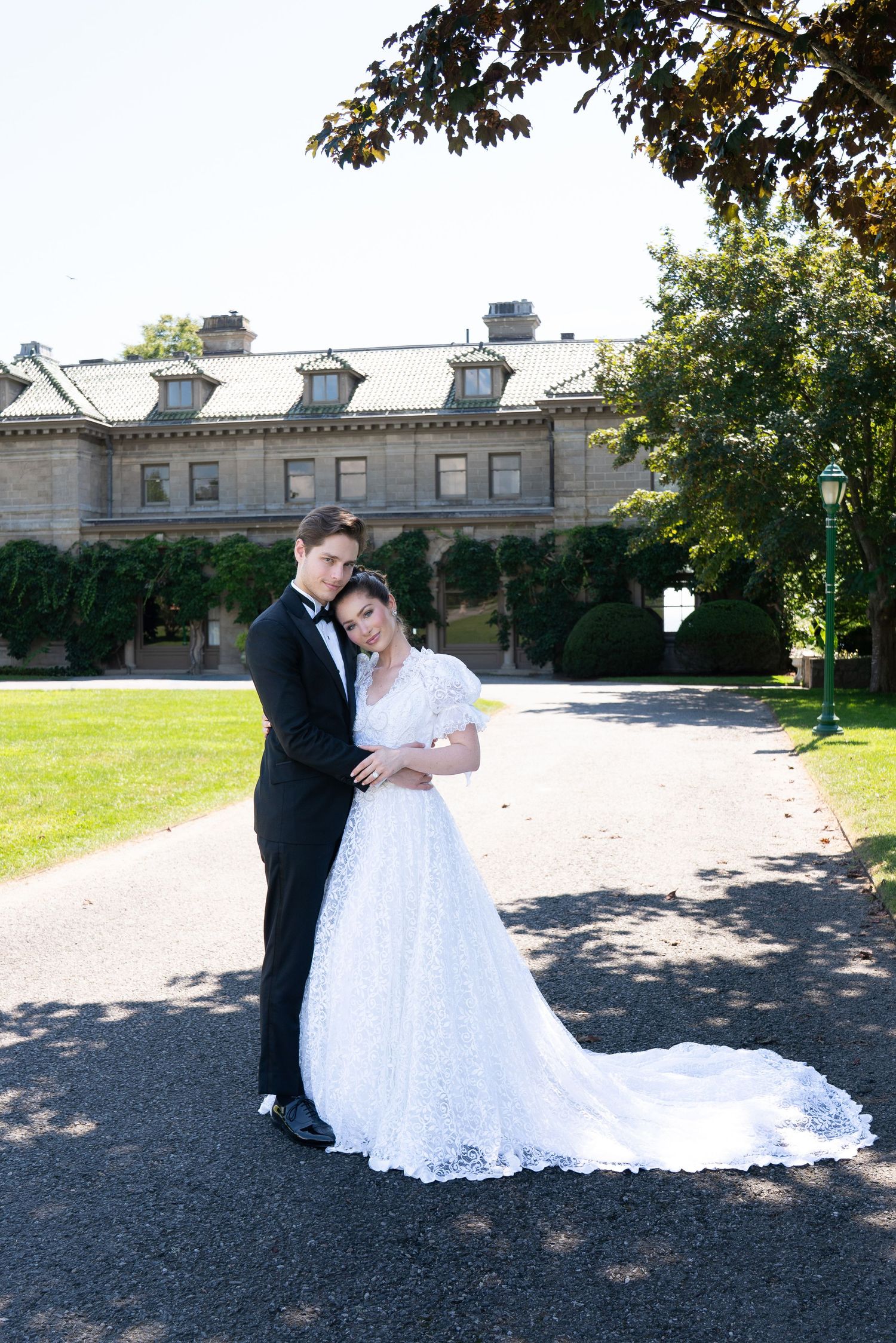 Newlyweds embrace on a sunlit path before a grand historic estate, bride in lace gown with train, groom in tuxedo.