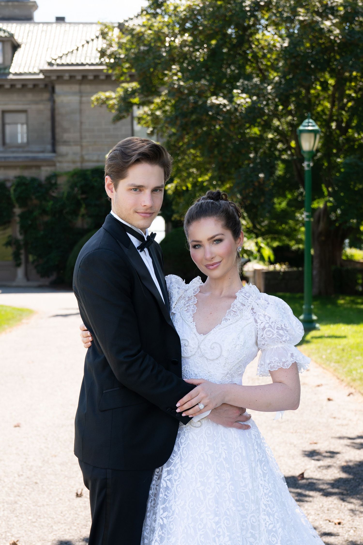 Elegant wedding couple embraces on a garden path near a historic estate, bride in lace gown, groom in black tuxedo.