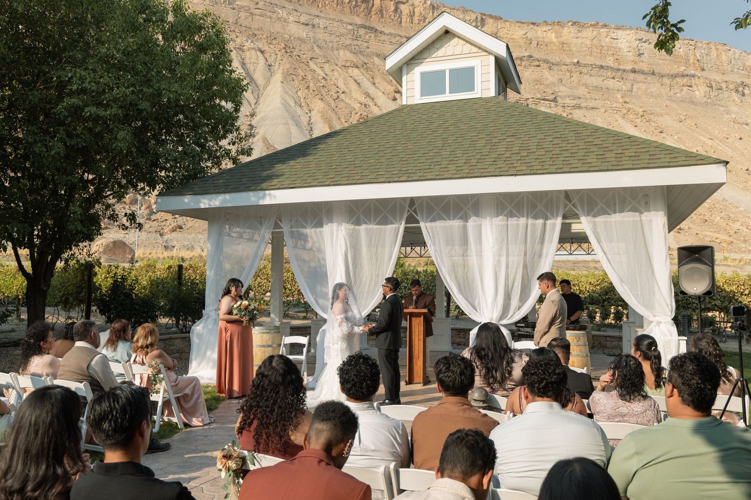 Outdoor wedding ceremony under a white gazebo with draped curtains, guests seated, desert cliffs in background.