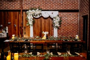 Elegant wedding head table with floral arch backdrop, greenery garland, candles, and black chairs in a rustic brick venue at Factory 51