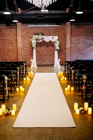 Elegant indoor wedding ceremony aisle with white runner, candles, dark chairs, floral arch, and exposed brick wall backdrop at Factory 51