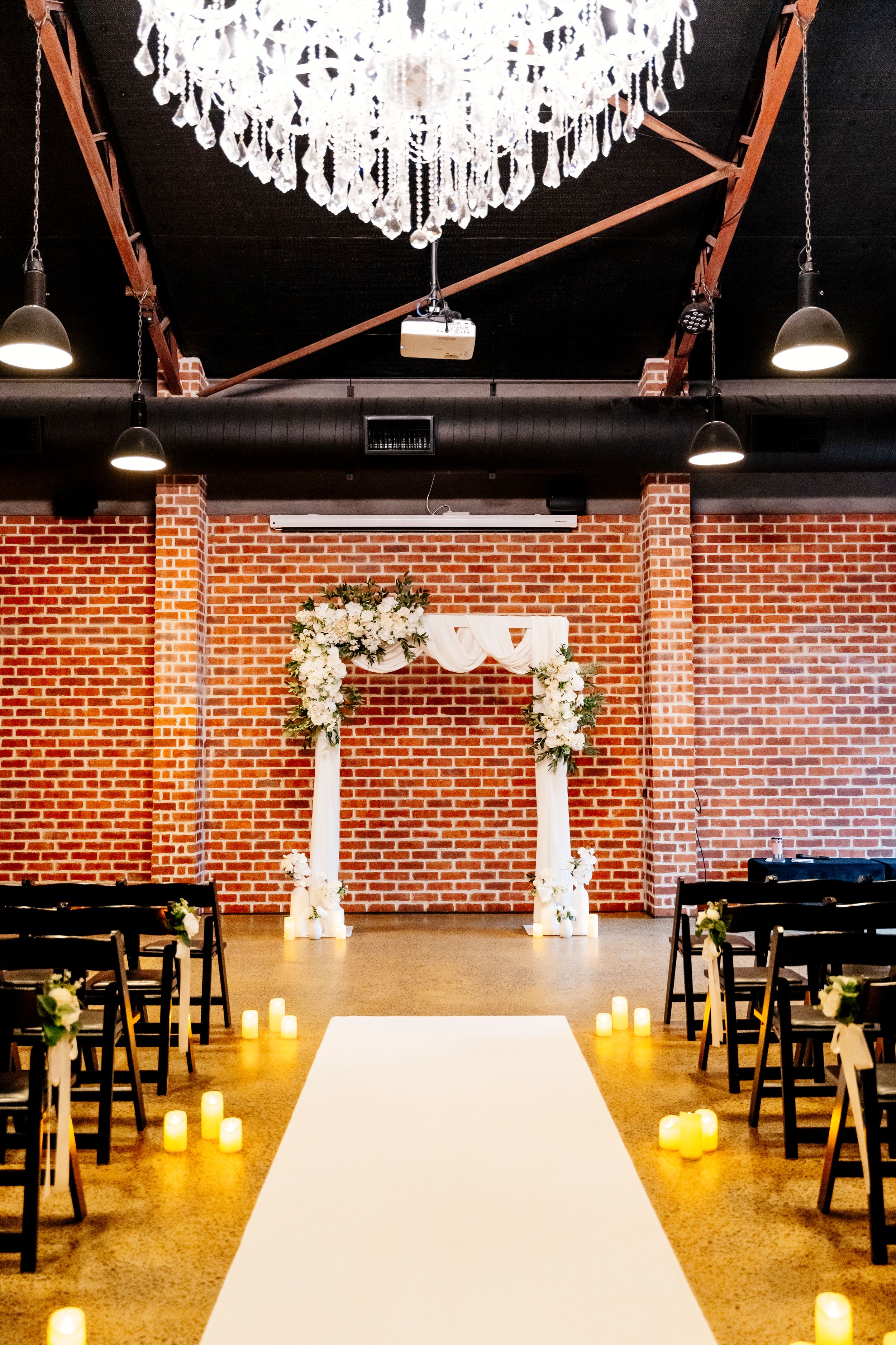 Elegant indoor wedding ceremony w/ floral arch, white aisle runner, candles, black chairs, and crystal chandelier in brick venue Factory 51
