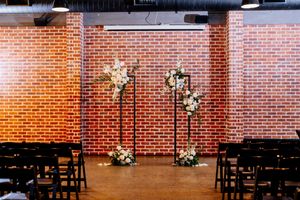 Elegant wedding ceremony setup with floral arrangements on black stand against a rustic red brick wall with dark wooden chairs at Factory 51