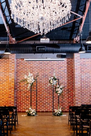 Industrial wedding venue with crystal chandelier, brick wall backdrop, floral arch stands, and dark wooden chairs for ceremony at Factory 51