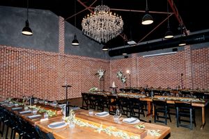 Elegant event dining room with crystal chandelier, brick walls, long wooden tables, and black chairs set for formal gathering at Factory 51