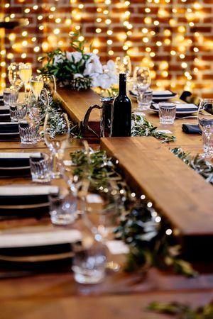 Elegant rustic wedding table setting with greenery garland, crystal glasses, black plates, and warm fairy lights backdrop at Factory 51
