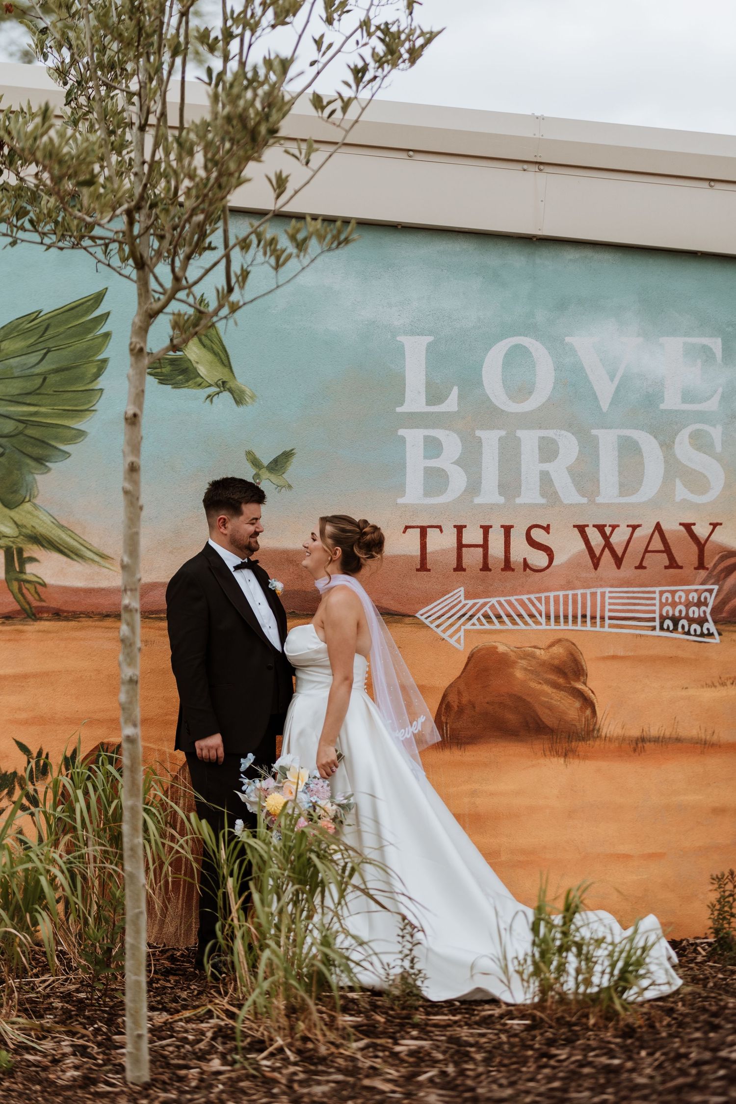 Newlywed couple sharing a romantic moment in front of a colorful Love Birds This Way mural outdoors.