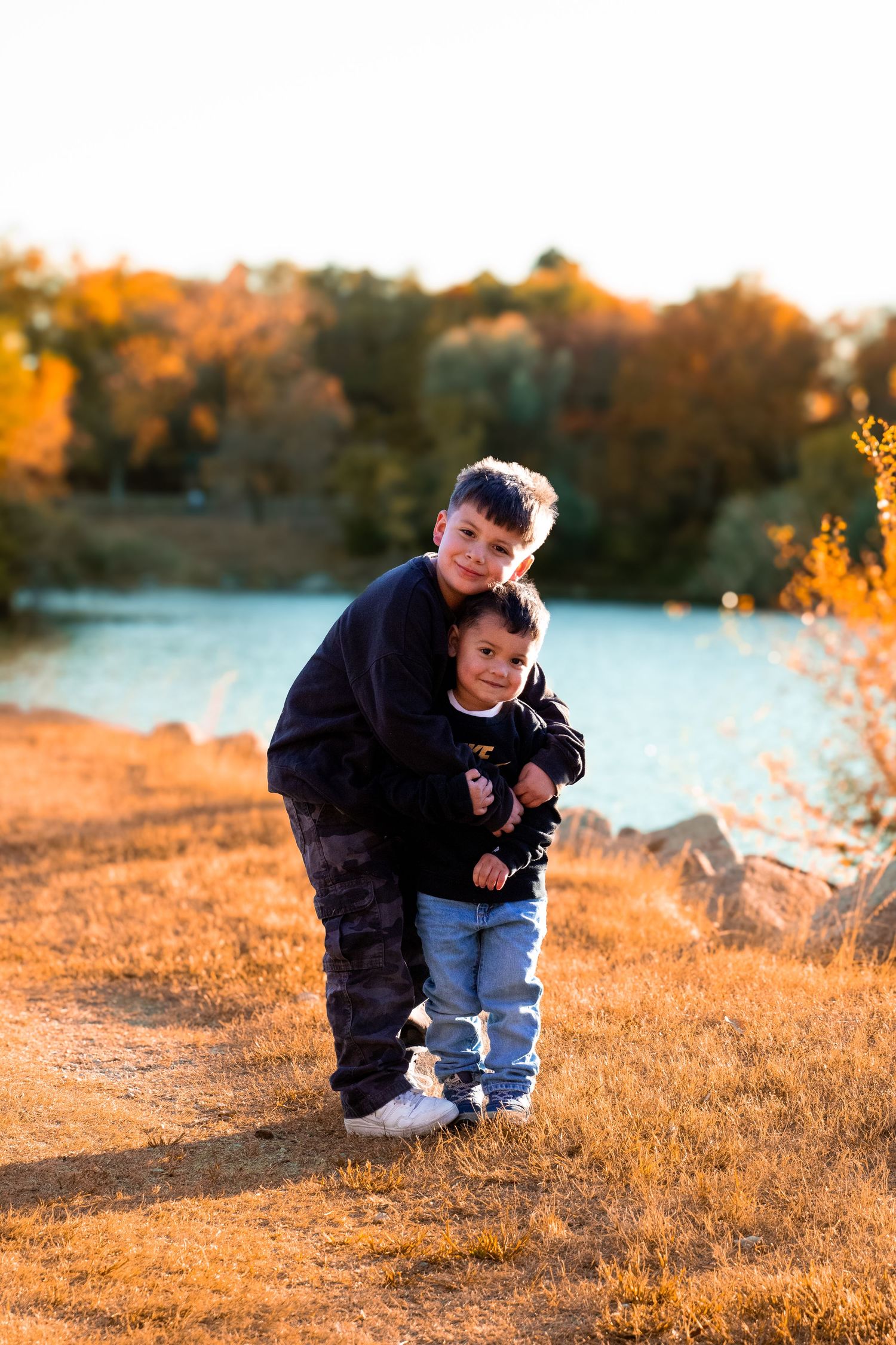 Two young boys hugging outdoors near a lake surrounded by colorful autumn foliage in warm golden light.