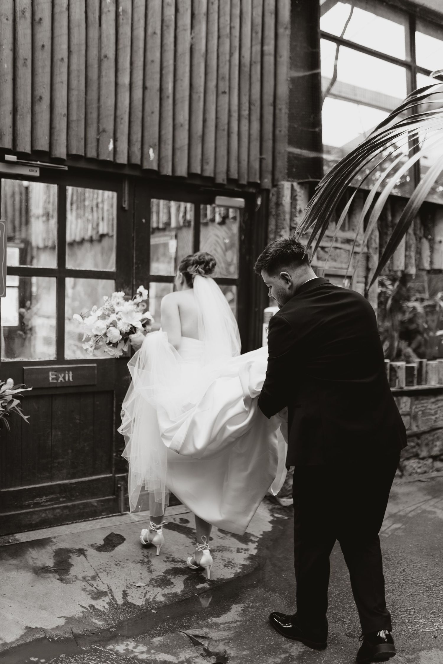 Groom in black suit helps bride in white gown with bouquet enter venue through exit door, black and white wedding photo.