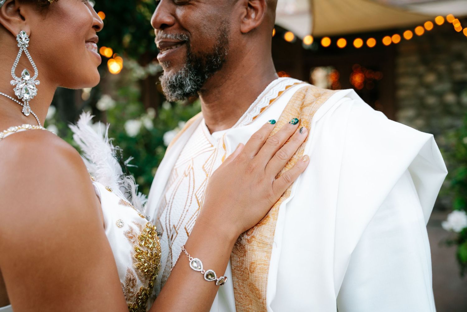 Couple in elegant African wedding attire sharing a tender moment outdoors with string lights in background.