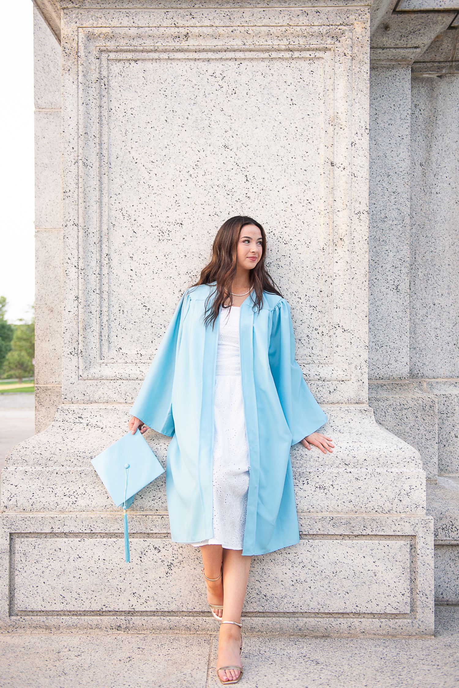 Young woman in light blue graduation gown and cap posing by stone columns outdoors.