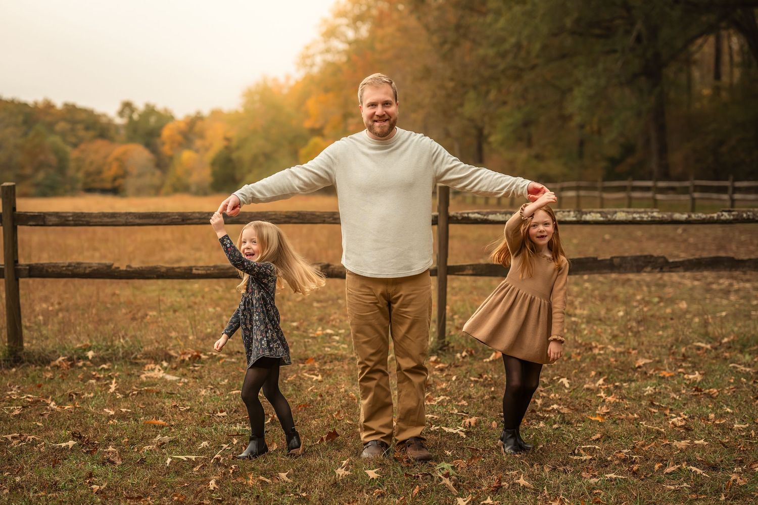A father holds hands with two young girls in a fall outdoor setting with wooden fence and autumn foliage.