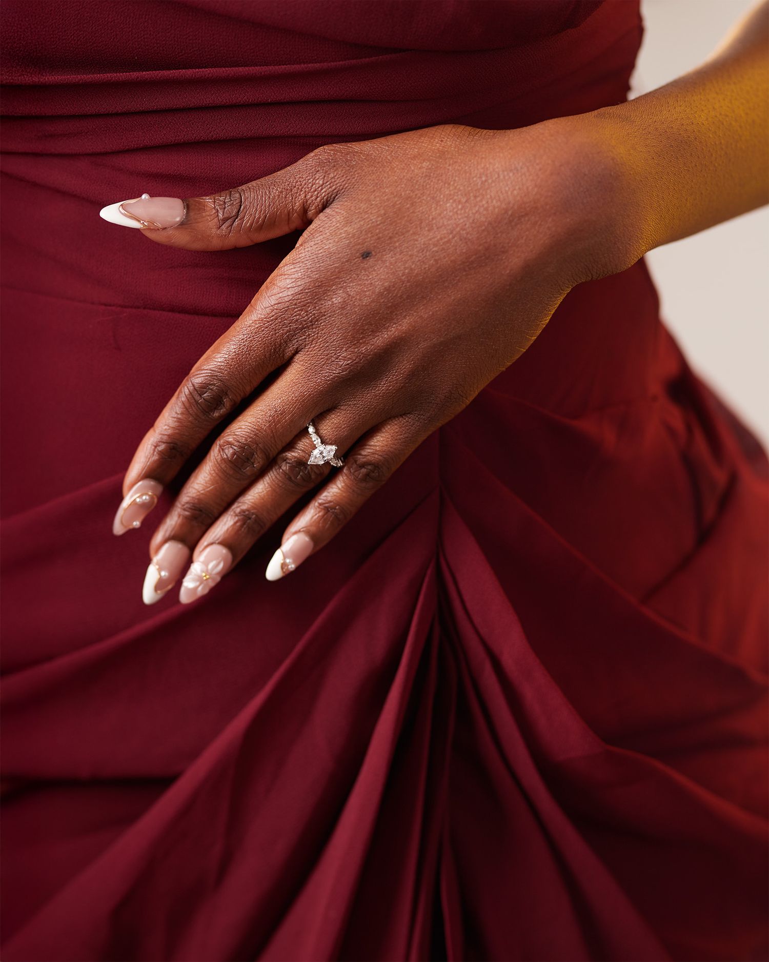 Close-up of a hand wearing a stunning diamond engagement ring, with white almond nails, against a red satin dress.