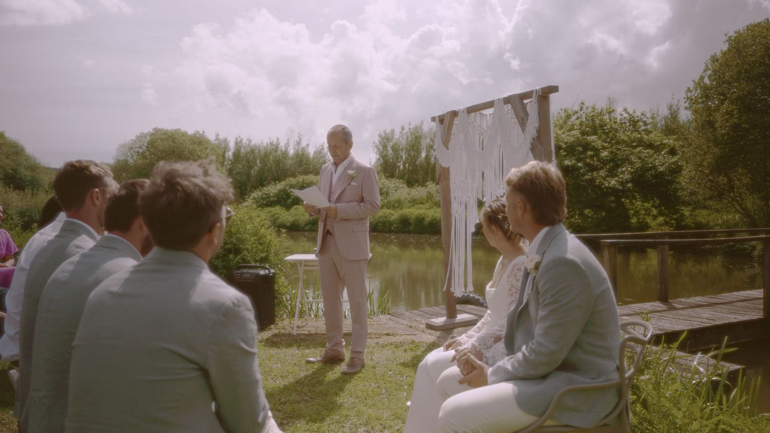Outdoor wedding ceremony by a pond with guests in blue suits and officiant in tan suit on a sunny day.