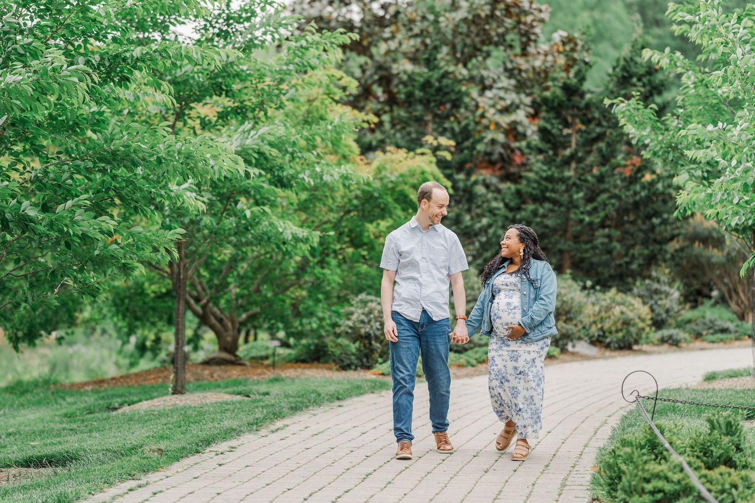 Couple holding hands and walking on a brick path in a lush green garden during an outdoor engagement session.