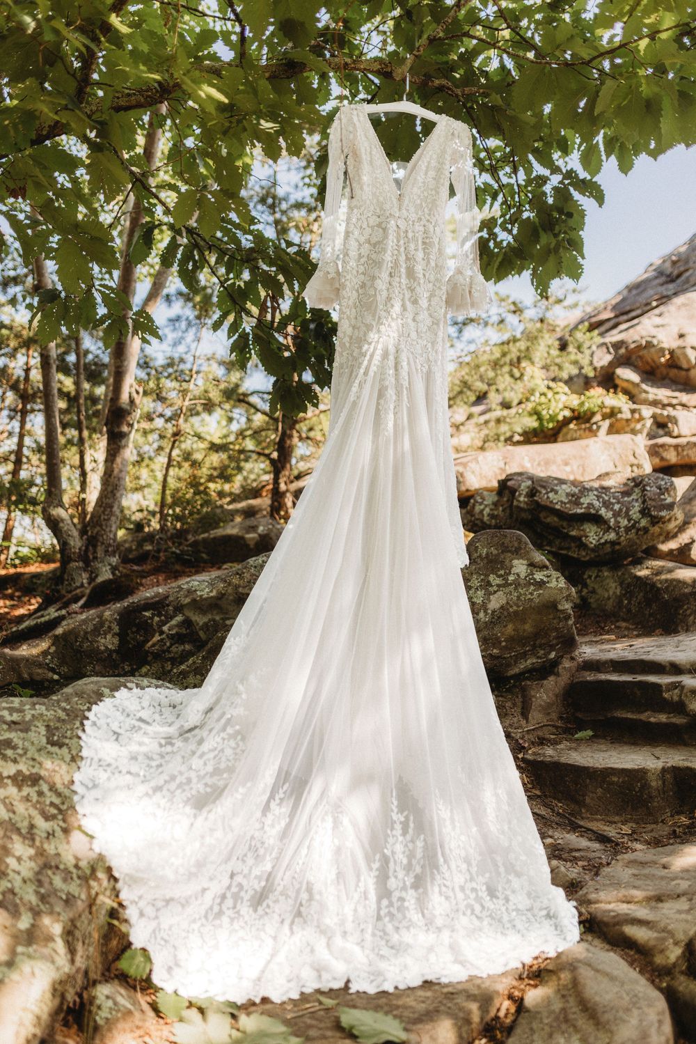 Elegant long-sleeve lace wedding dress with train hanging from tree branch over stone steps.