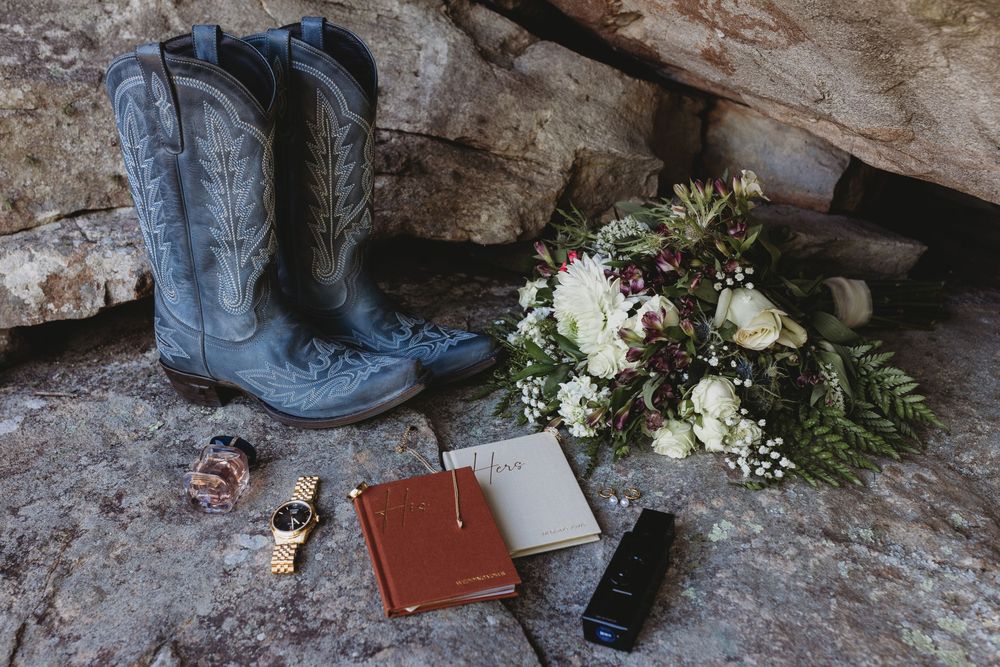 Wedding flat lay with blue cowboy boots, bouquet, vow books, watch, and accessories on rocks.