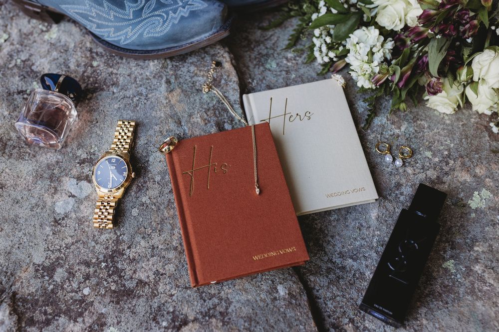 Wedding detail flat lay featuring His and Hers vow books, gold watch, jewelry, and bouquet.