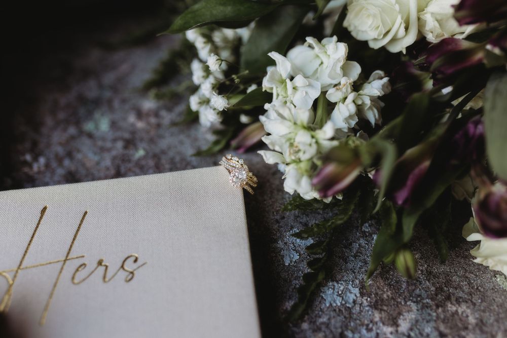 Wedding ring resting on bouquet of white flowers beside a gold foil Hers vow book.