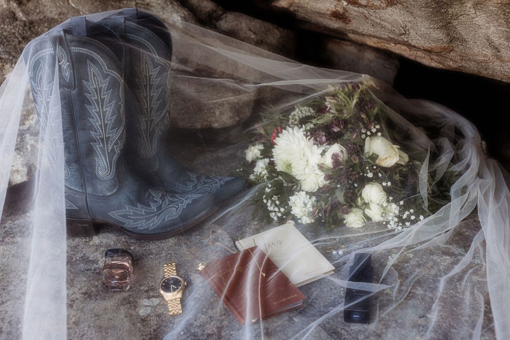Bridal flat lay with cowboy boots, floral bouquet, rings, and veil on rocky surface.